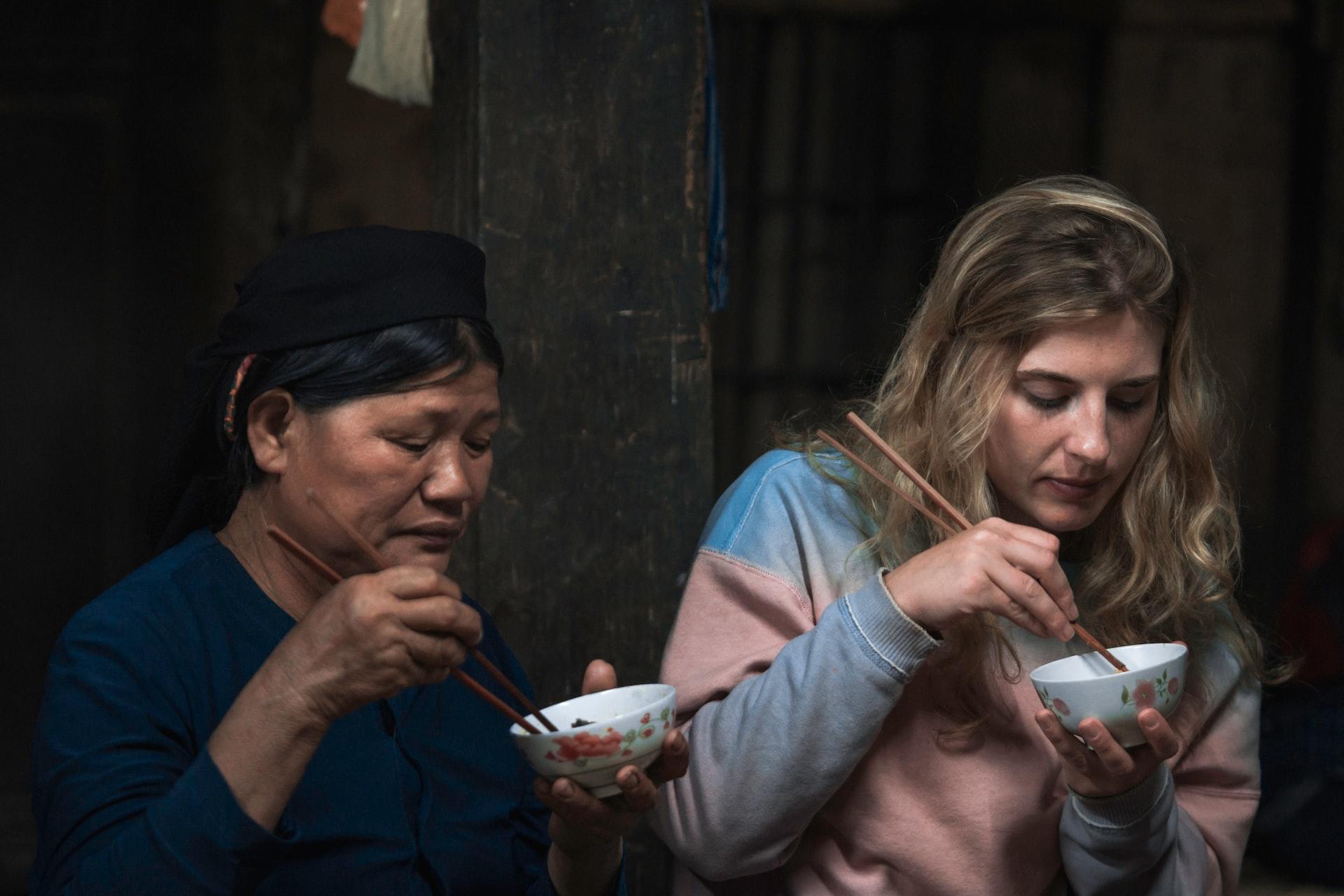 A blond woman and an Asian woman sitting side by side eating out of bowls with chopsticks.