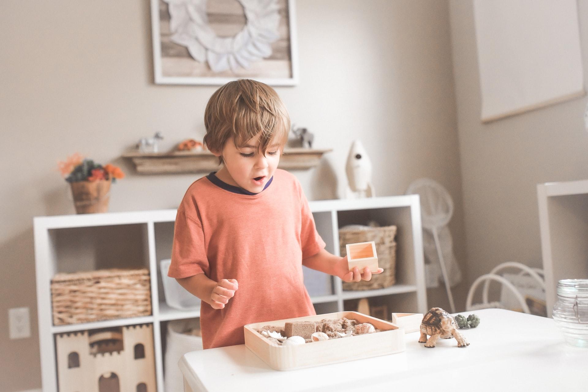 A boy in an orange top stands in front of a low-level storage shelf and marvels over a wooden box of materials. 