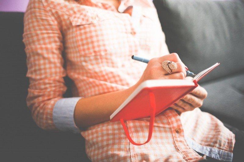 A person in an orange and white checked shirt reclines against a dark sofa to write in a red hard-bound notebook.