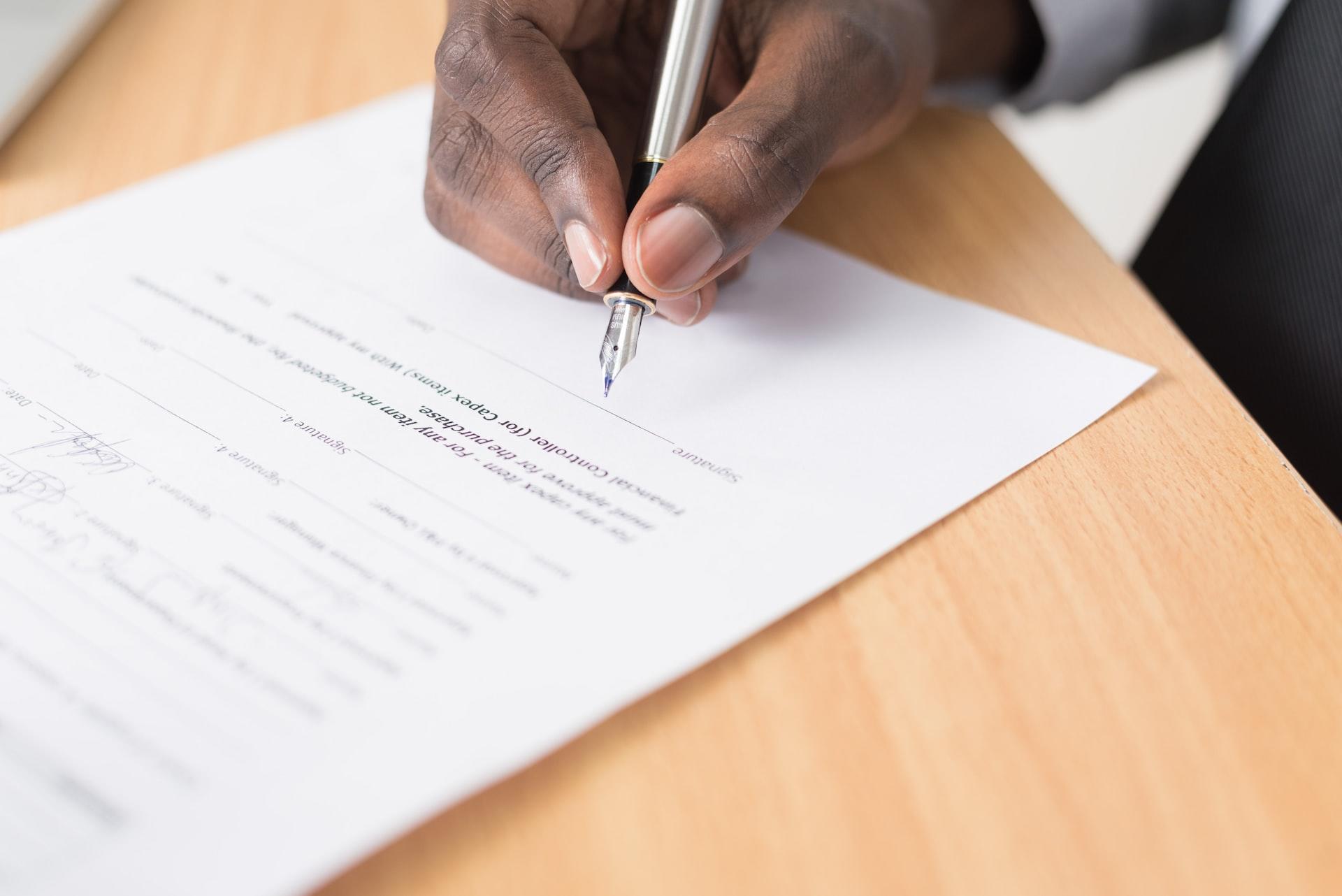 A hand holding a pen preparing to write on an exam paper placed on a blond wood desk