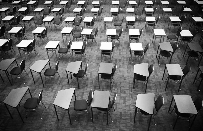 A greyscale image of an exam centre room, with tables and chairs lined up in rows on a carpeted floor but no students yet present. 