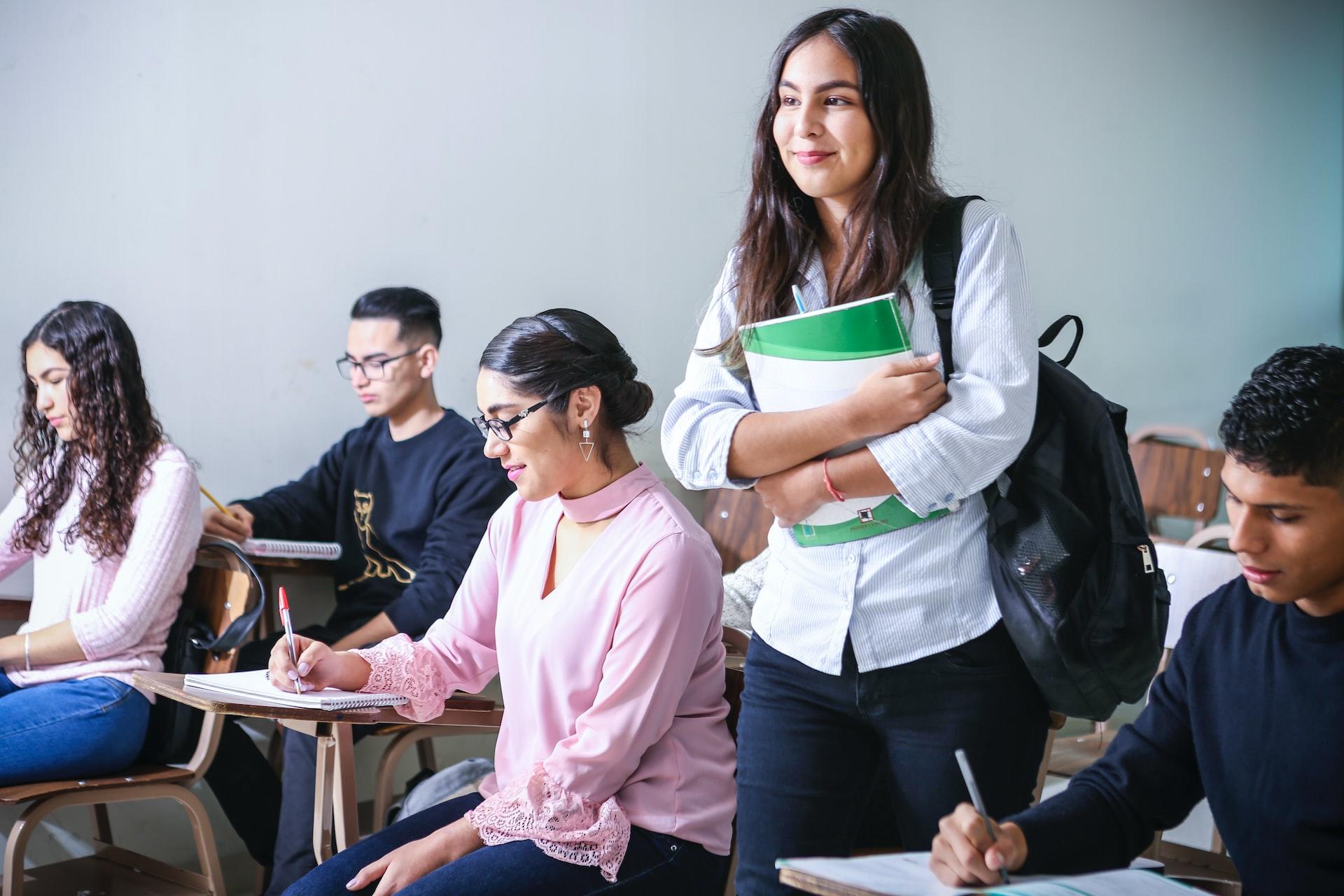 A class full of seated students with one young woman standing, with a backpack over one shoulder and a green-covered book in her arms.