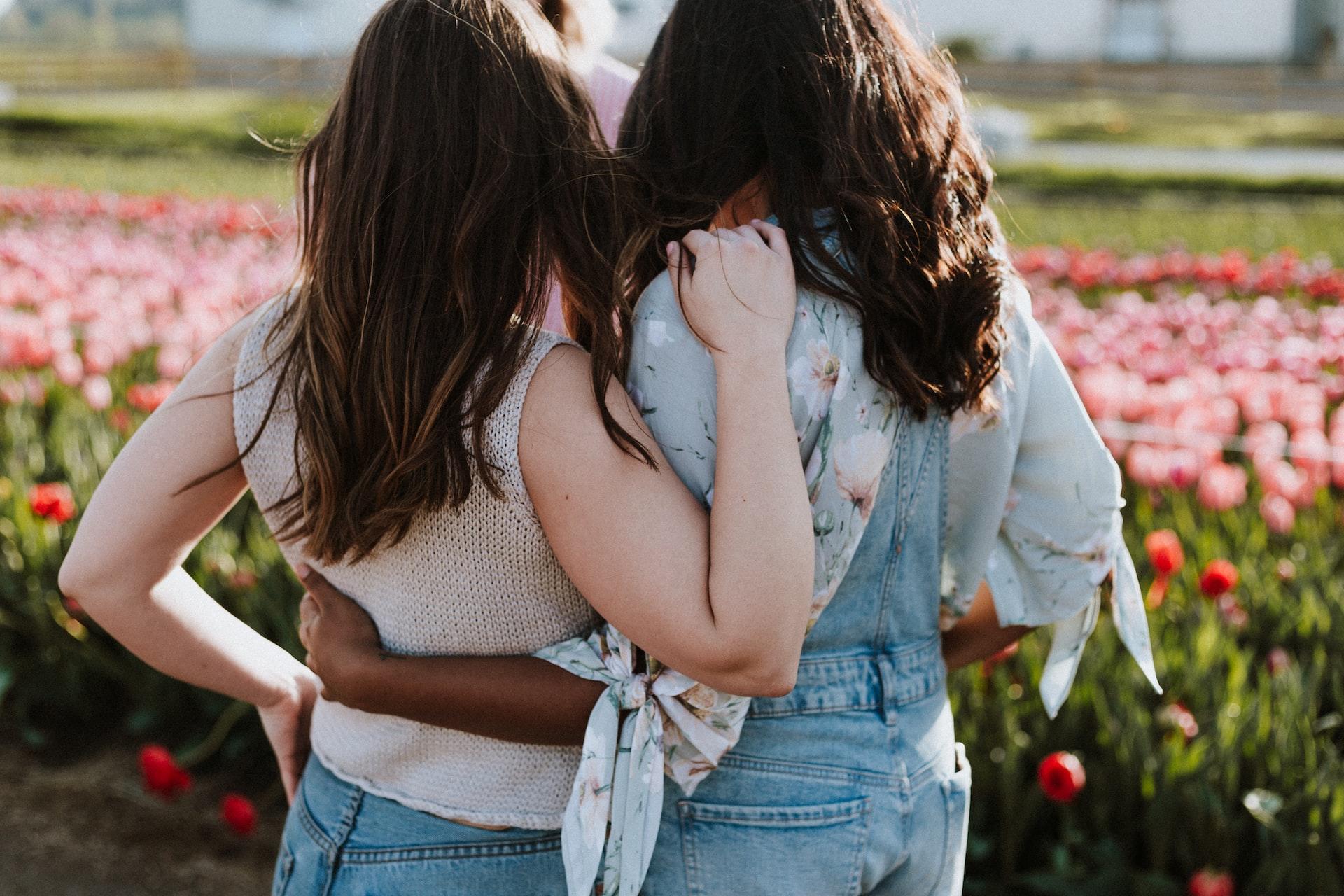 Two women with long hair standing arm in arm in front of a field of flowers