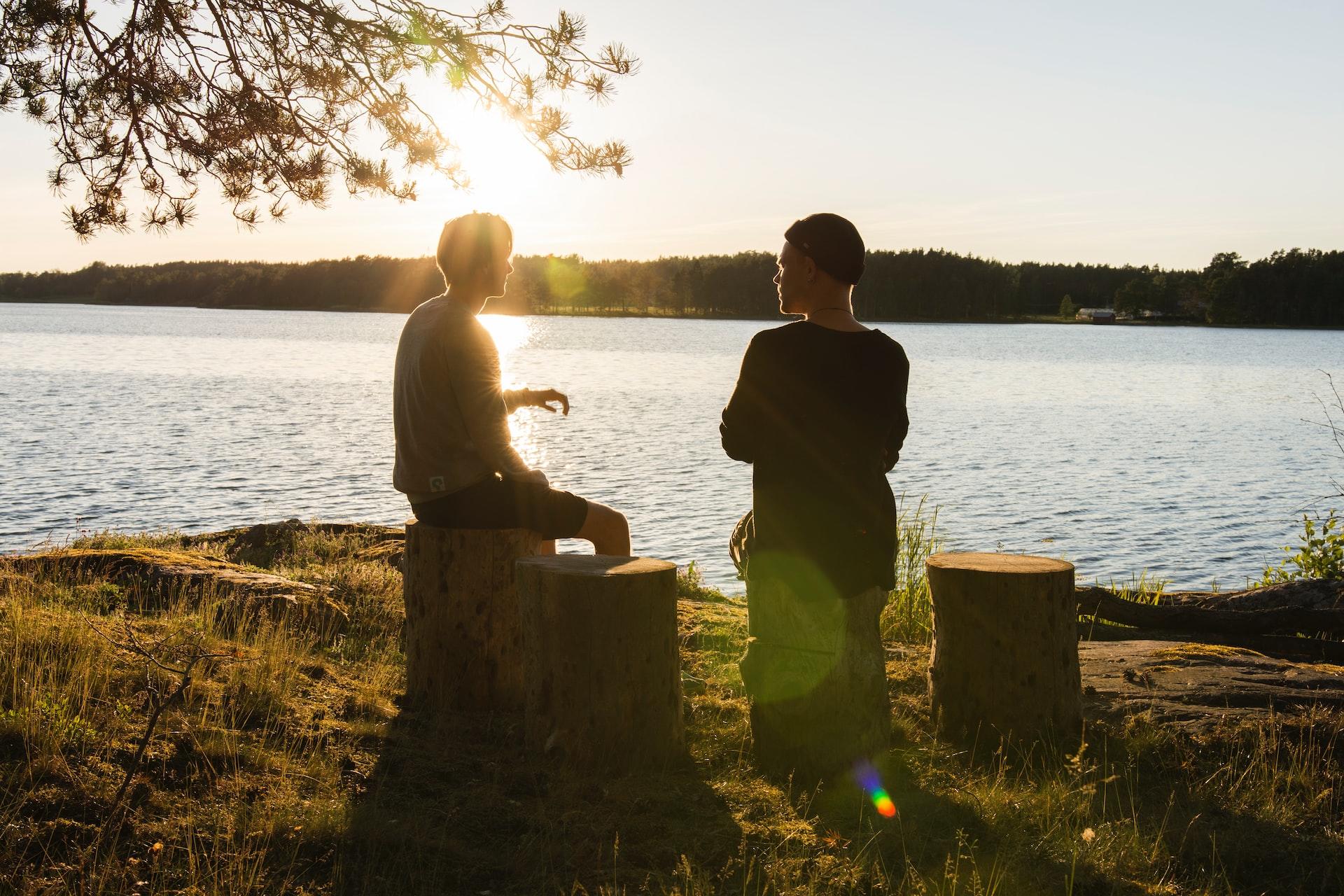 Two people sitting on tree stumps by a body of water at sunset, slightly turned from one another but apparently talking. 