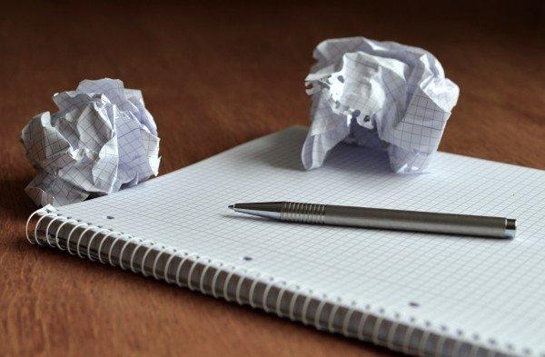 A spiral notebook open to a blank page with a silver-coloured pen laying on it rests on a wooden surface, with two sheets of paper crumpled up next to it. 