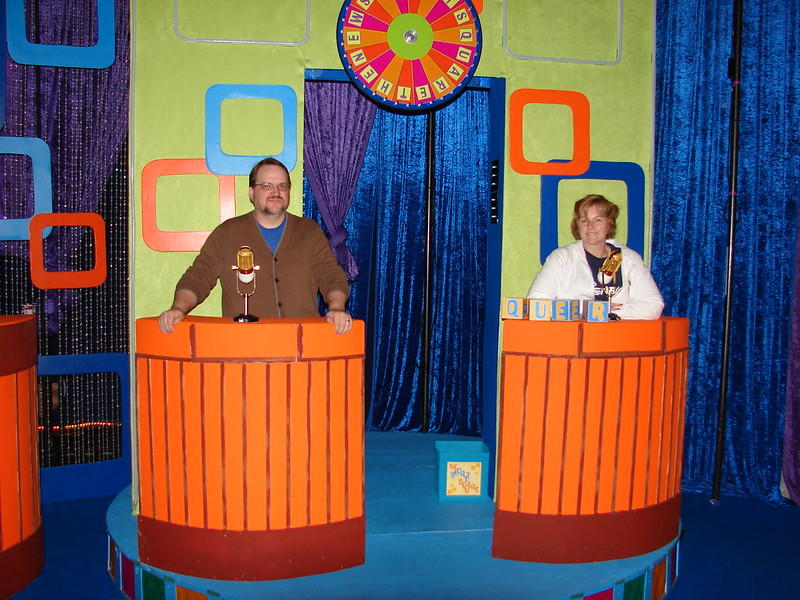 A game show stage set-up with orange contestant podiums set on a light blue platform against a lime green backdrop framed with sparkly blue and purple drapes. 