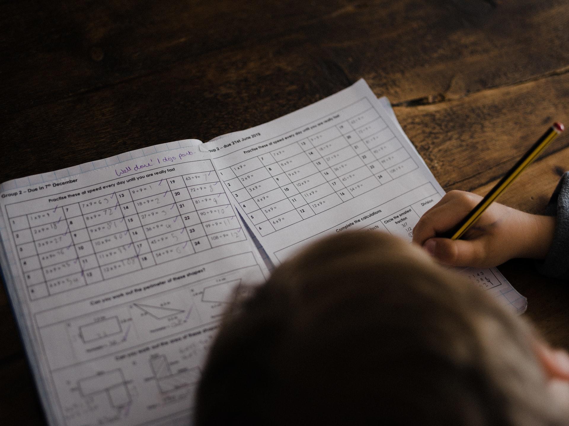 A person sitting at a wooden table writing on a collection of maths worksheets