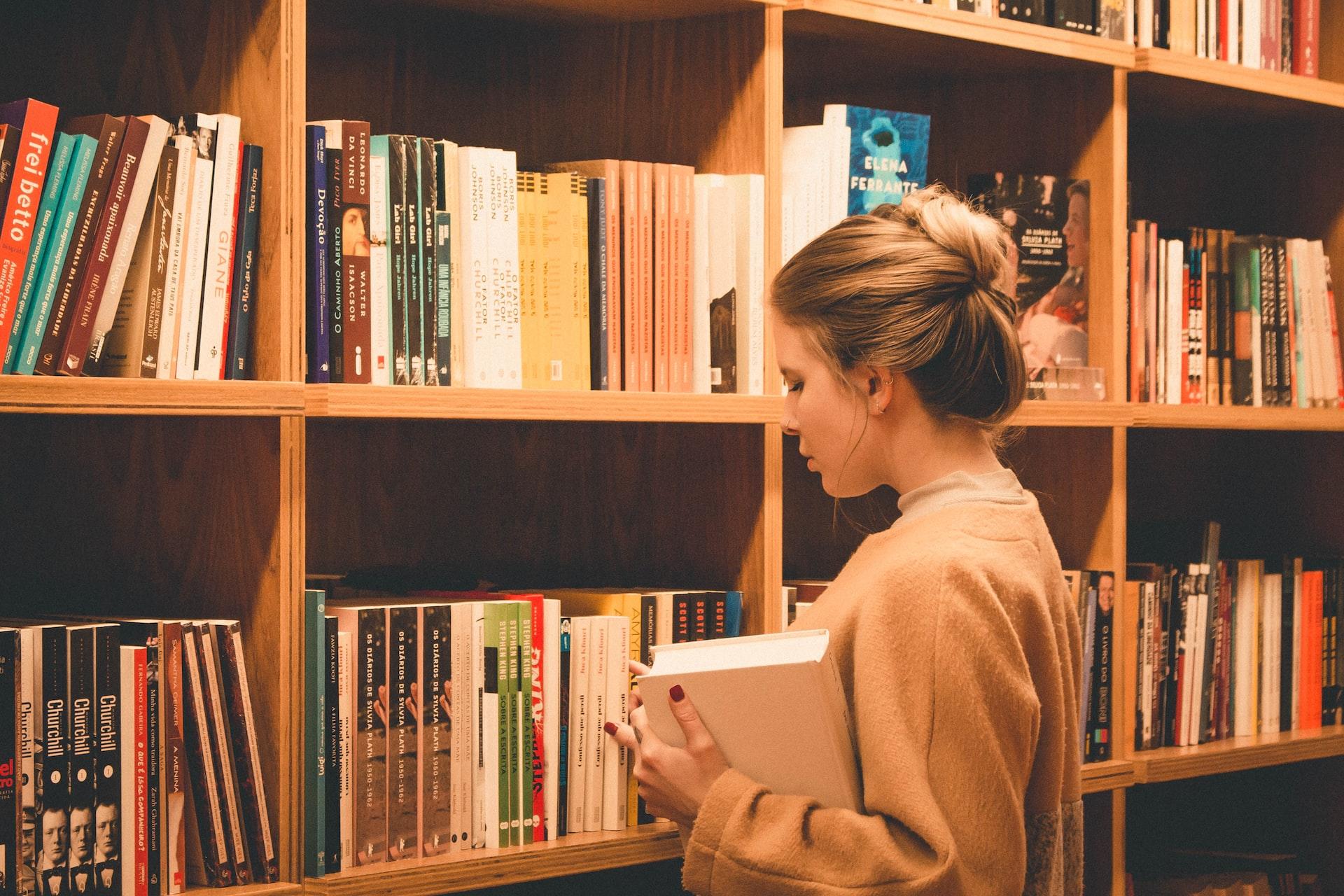 A woman in a tan jumper cradles a thick book in her left arm while looking at a full bookshelf with a worried expression