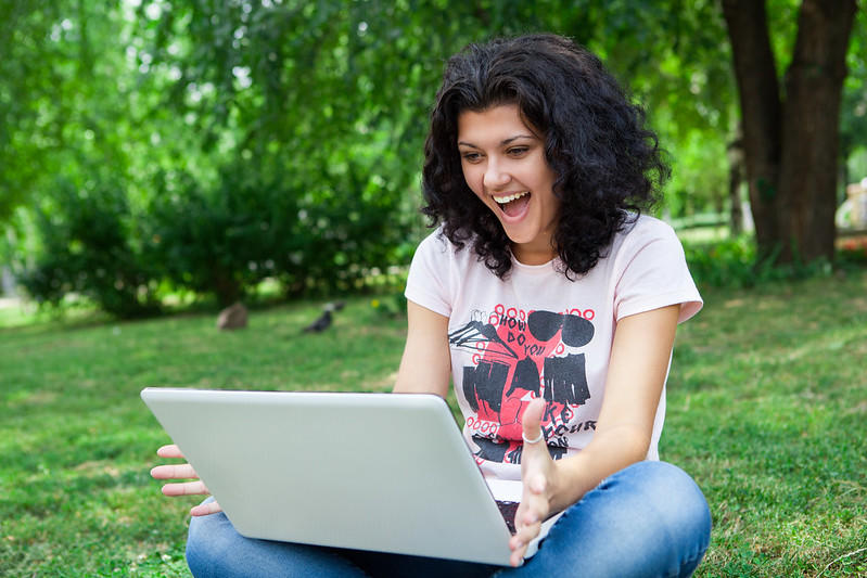A young woman with dark hair wearing blue jeans and a white shirt sits cross-legged in the grass with an open laptop balanced on her knees.