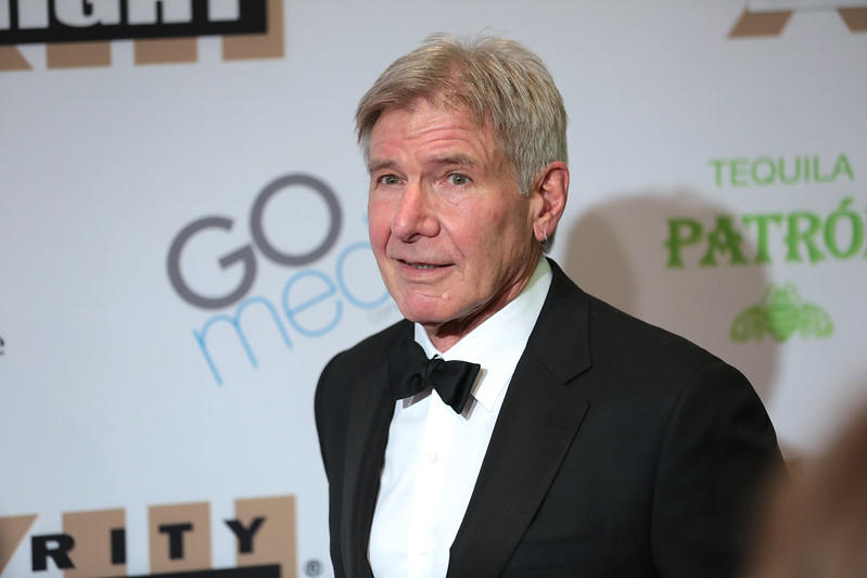 Actor Harrison Ford, wearing a black tuxedo with a white shirt and bowtie, standing in front of a background full of brand names