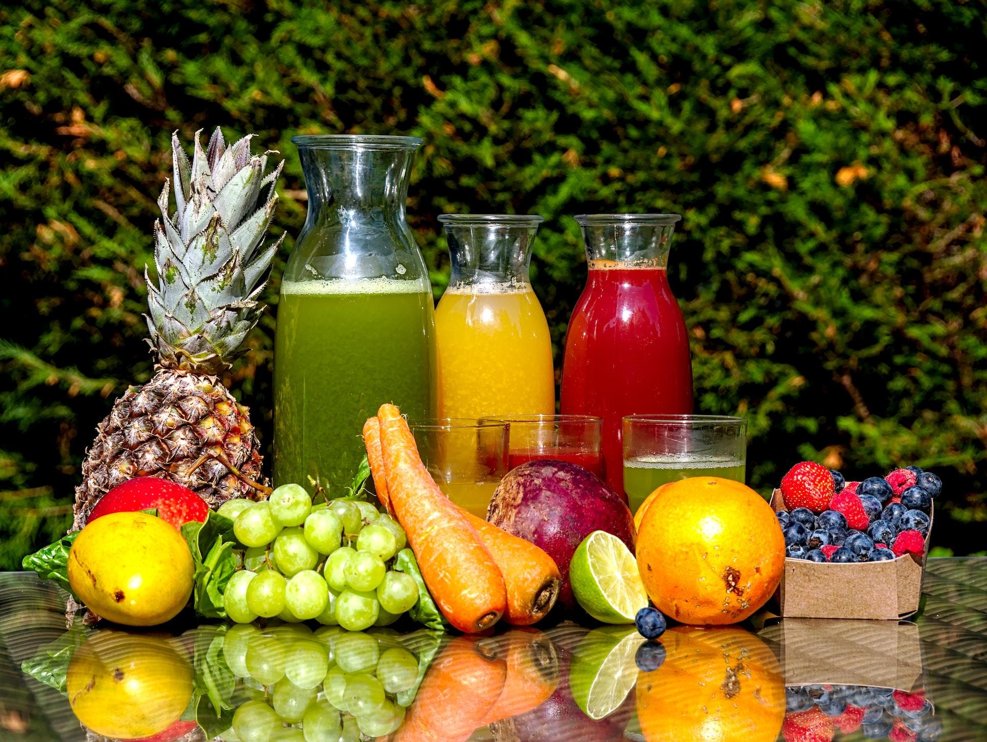 Three bottles of fruit juice, one green, one orange and one red, behind an array of fresh fruits and vegetables