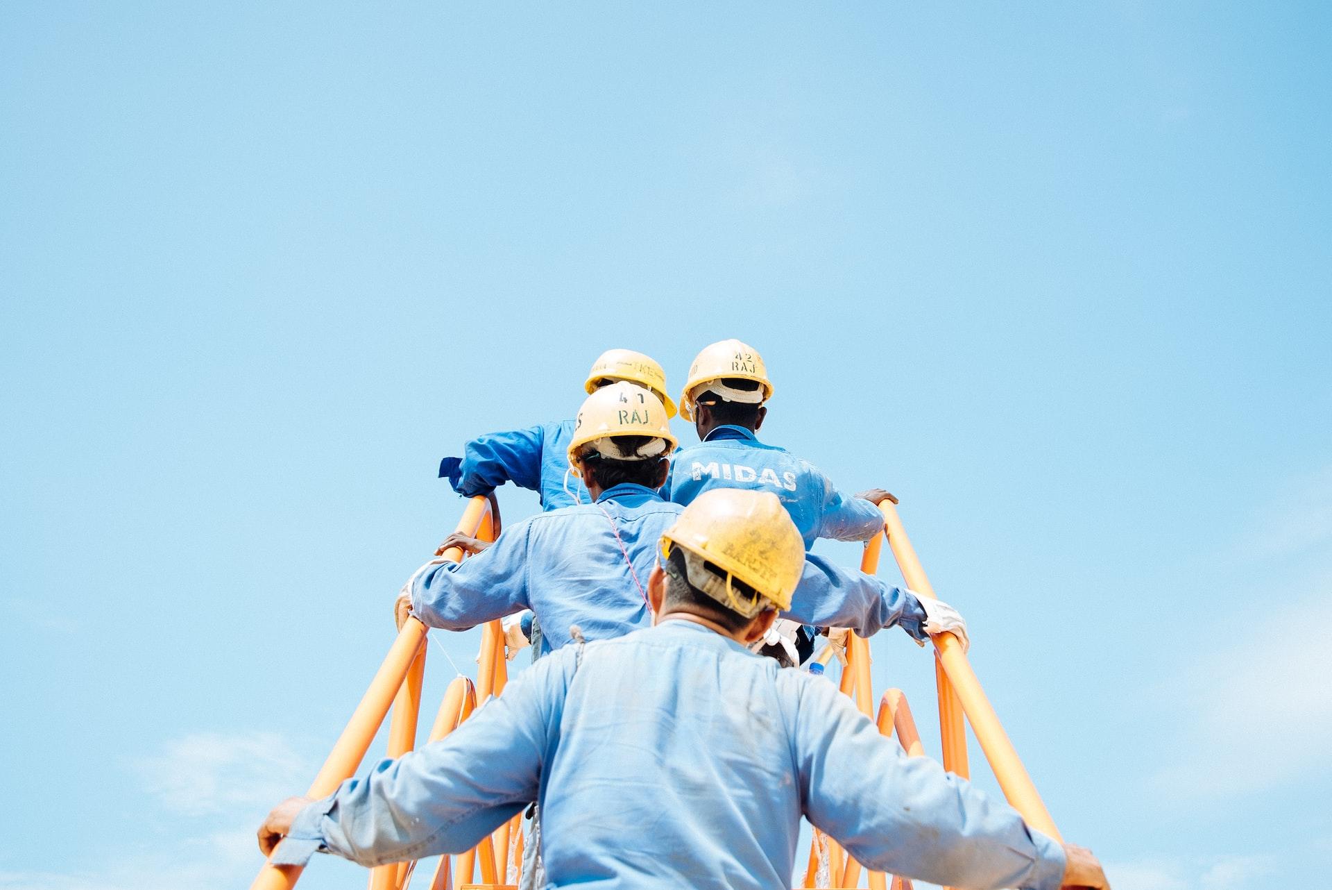 A line of workers, all wearing light blue shirts and yellow hard hats, climbing a ladder with yellow guardrails