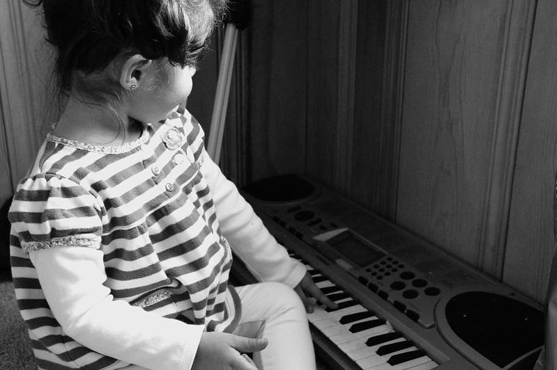 A black and white picture of a toddler in a striped dress over long sleeves picking out notes on an electronic keyboard. 