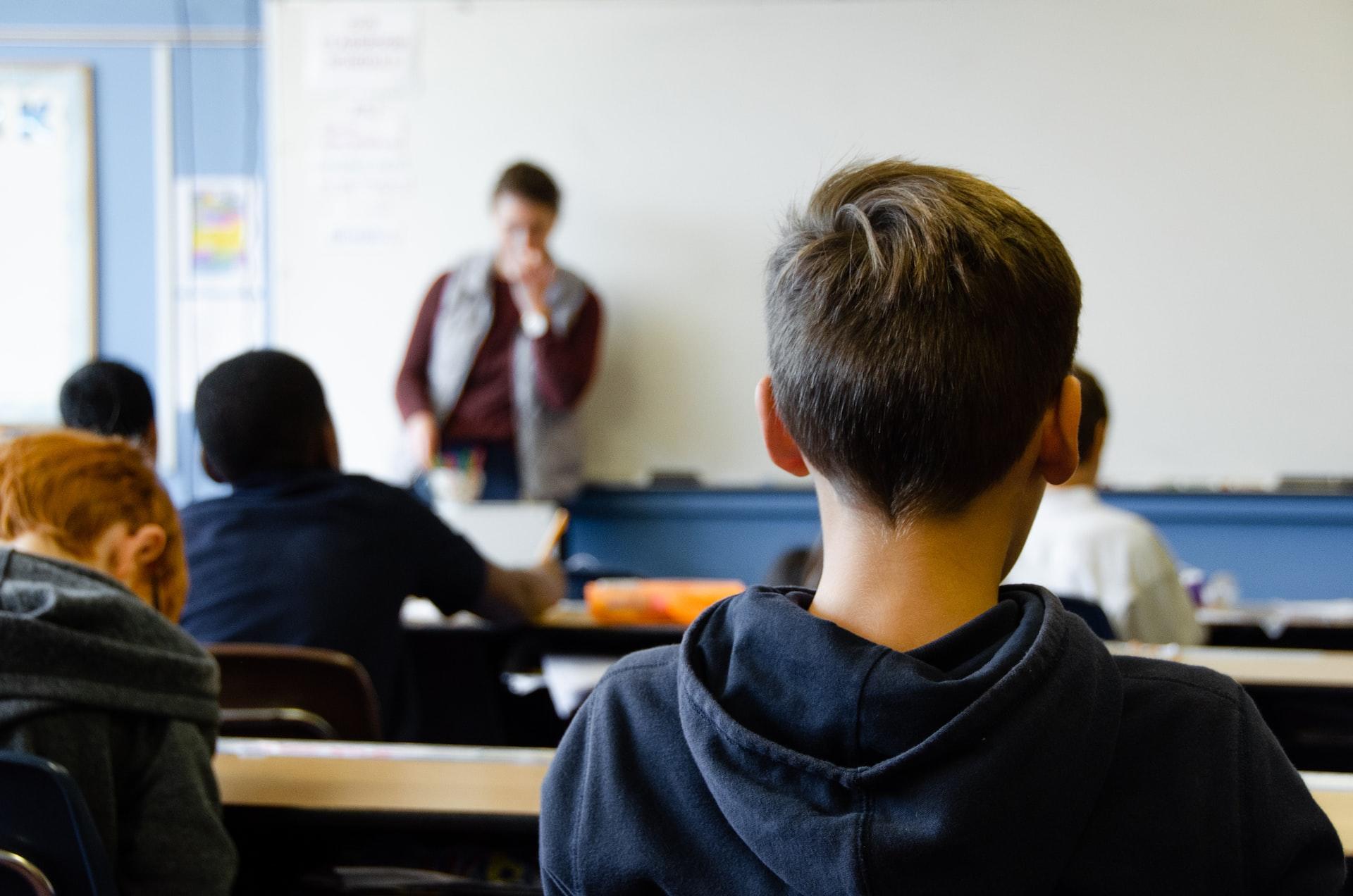 A boy in a blue hoodie sitting in a classroom looking at the teacher