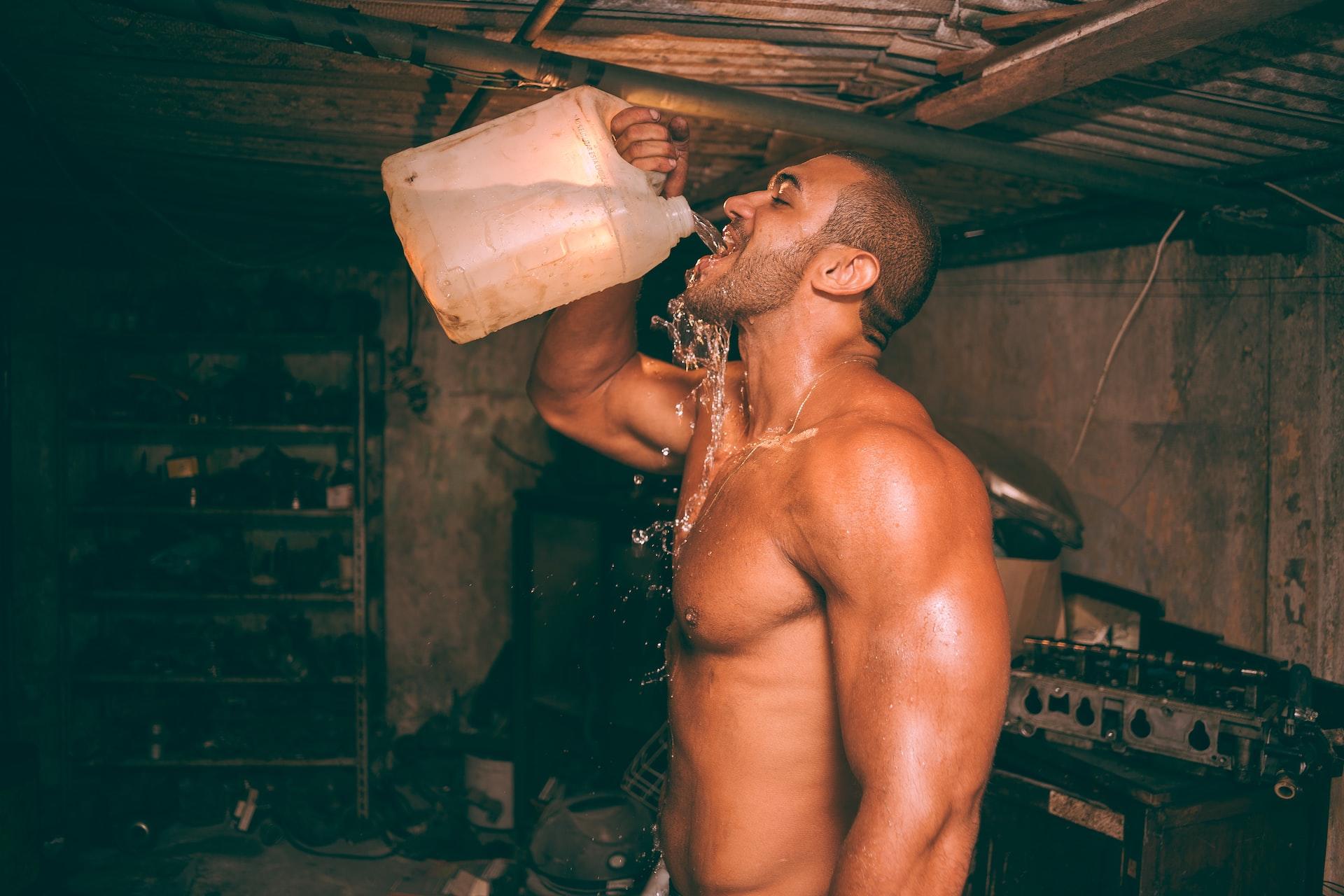 A shaven-head, shirtless man stands in a darkened shed drinking water from a 5-litre white plastic jug. 