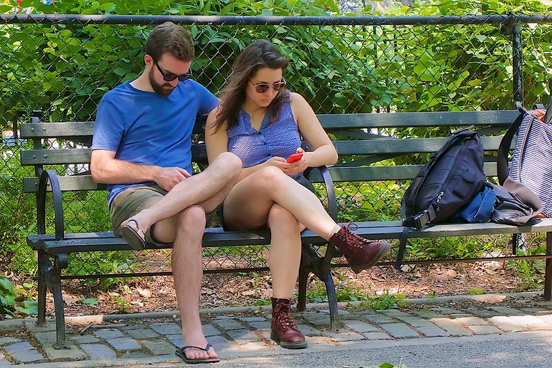 A man and a woman, both wearing blue shirts, shorts and sunglasses, sit close to one another on a park bench looking at their phones. Their backpacks are at the other end of the bench.
