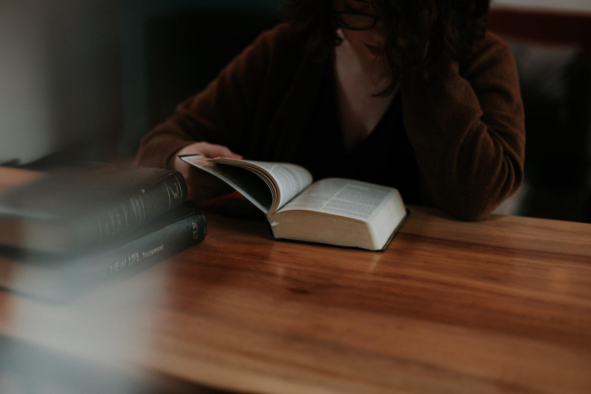 A person sitting at a wooden table reading a bible with two other hard-bound books nearby