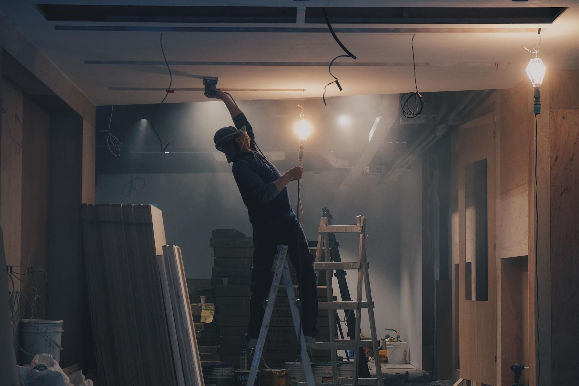 A finishing worker stands on a ladder to apply a coat of paint on the ceiling in a dimly-let, unfinished interior space