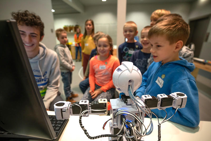 A group of students observes a robotics demonstration, focusing on the computer display while the small robot stands to the side with its arms outstretched. 