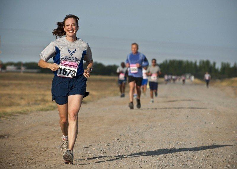 A woman wearing blue shorts and marathon number on a grey tee-shirt smiles as runs on a dirt path ahead of other runners.