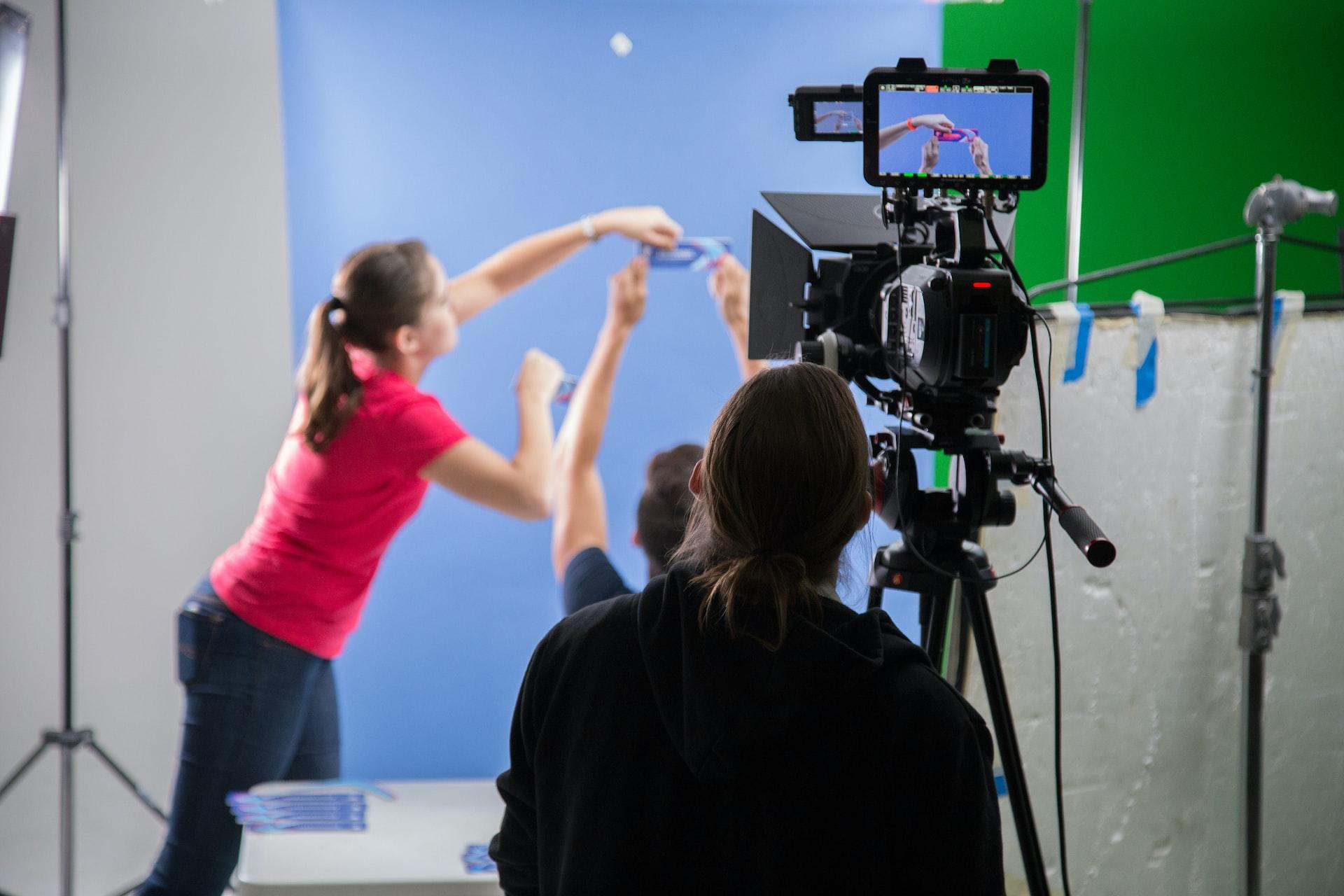 A woman in a bright pink shirt leans forward with her arms raised to touch up an actor's makeup while the camera points directly at them