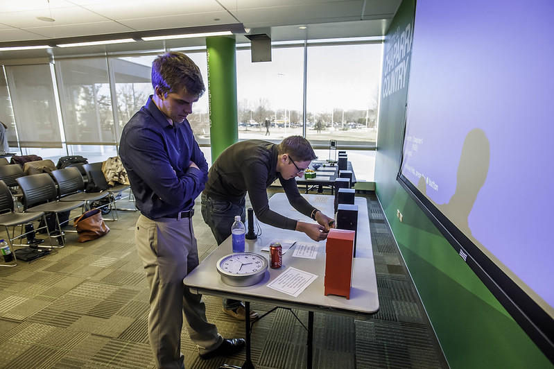 Two men wearing business casual attire stand near a folding table upon which rests a series of upright blocks, all but one coloured red. 