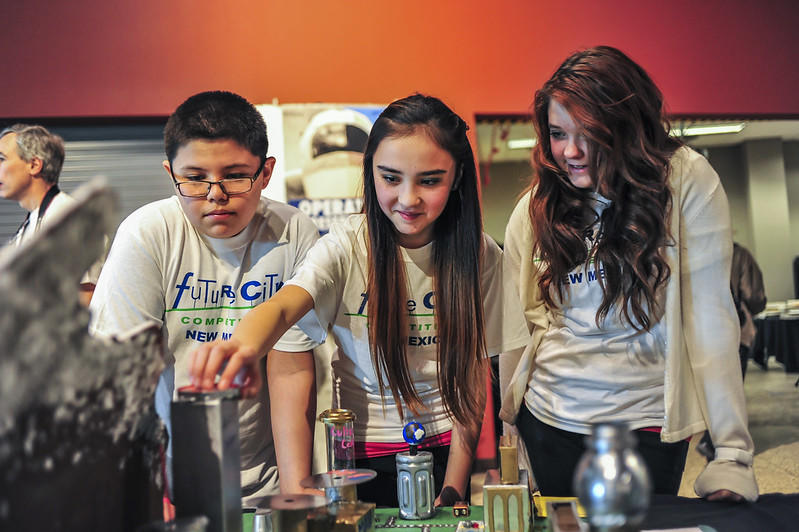 Three students wearing white tee-shirts with blue and green writing on them adjust their science fair fair project in a room with orange walls. 