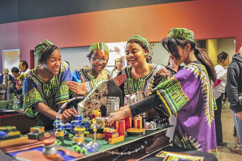 A group of young women wearing green, purple and blue dashikis with matching hats smile as they play with their science invention in a room with orange walls. 