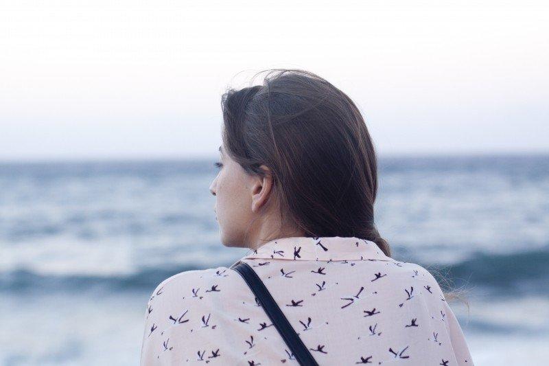 A woman wearing a white and blue patterned shirt, with her purse strap crossed over her back, gazes out onto the ocean.