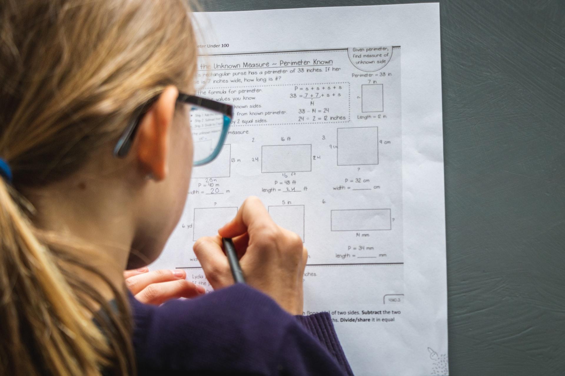 A blonde girl with glasses, seen from behind, as she works on maths problems written of a sheet of paper.