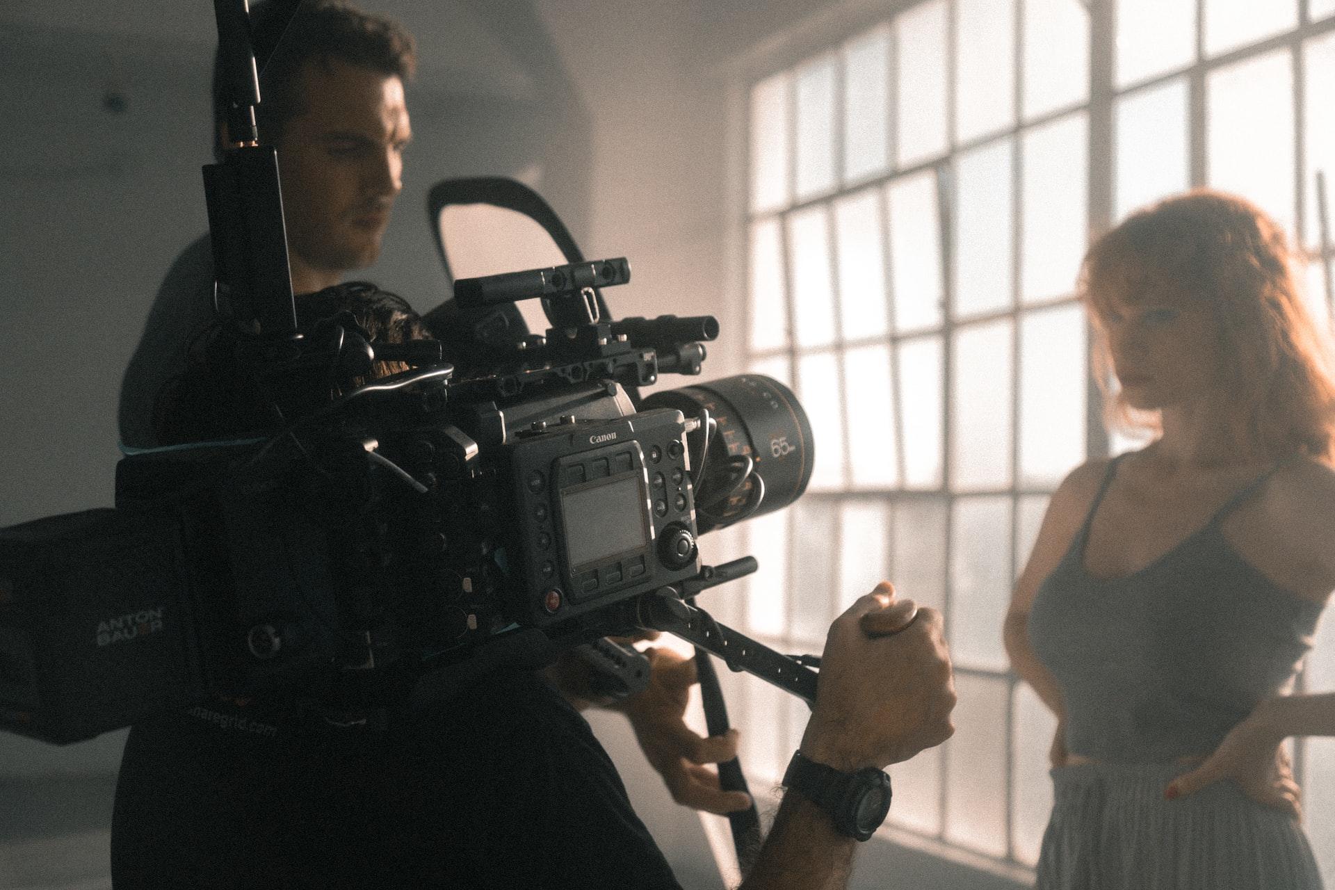 A woman in a tank top standing in front of a bank of windows while a technician points a camera at her and another actor faces her