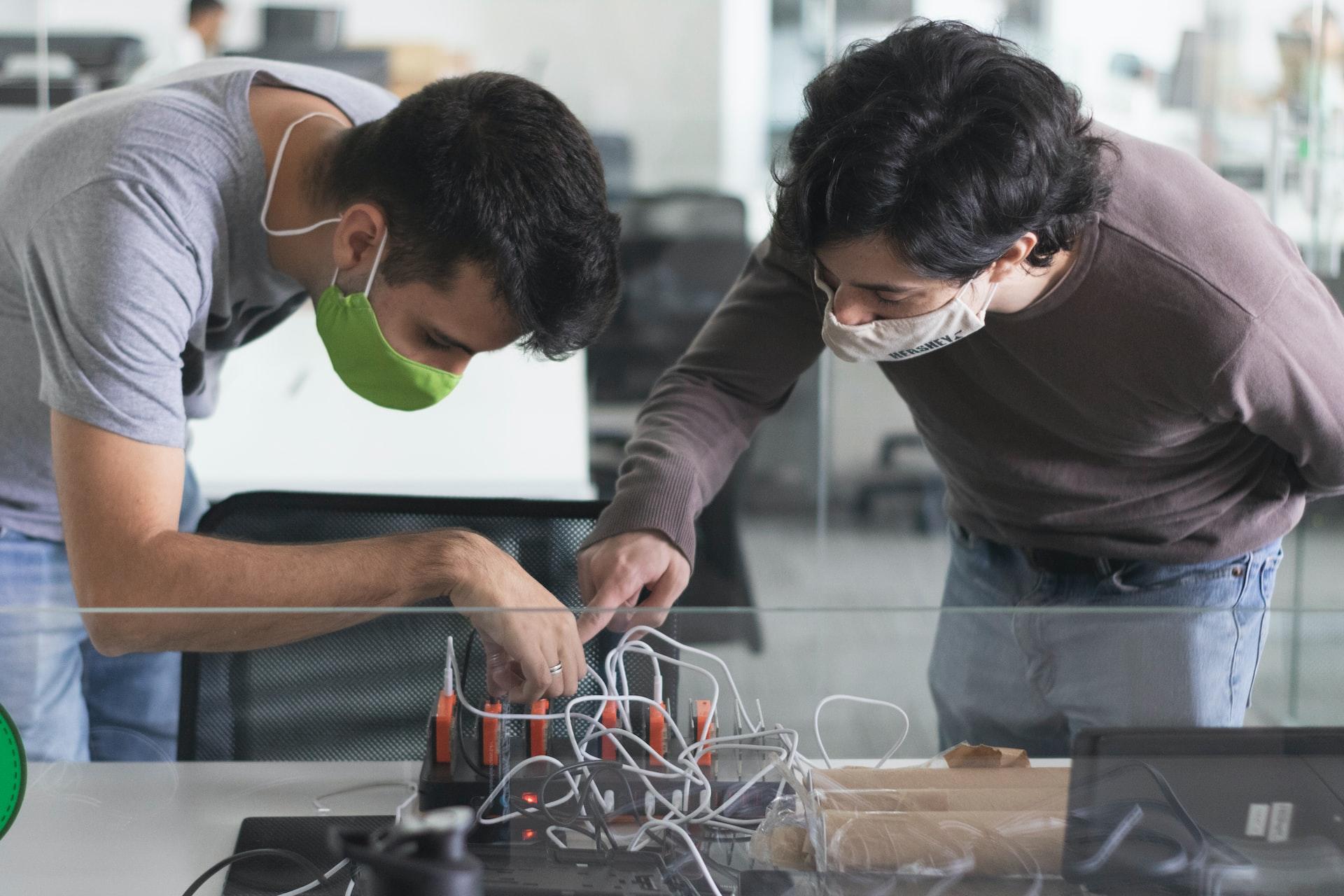 A student wearing a green face mask gets advice from his teacher wearing a brown jumper as they look at his robotic design