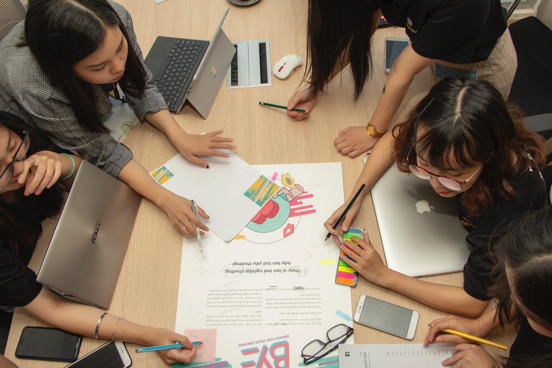A group of students discuss a project they've printed out to place in the middle of the table. Each has a laptop in front of them but they do not use it, focusing on the printed poster instead.