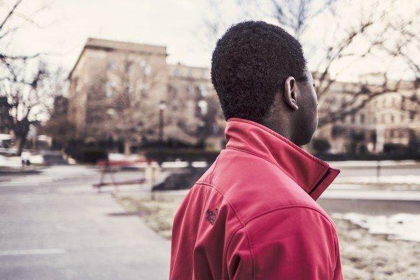 A youth wearing a red zip-up jacket with the collar turned up faces away from the camera, looking out over a park that's barren in winter. 