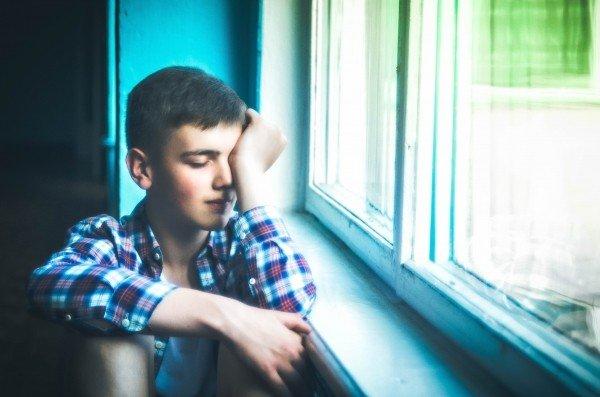 A boy in a plaid shirt sits by the window in a room with blue walls, with one hand covered half of his face and his eyes closed. 