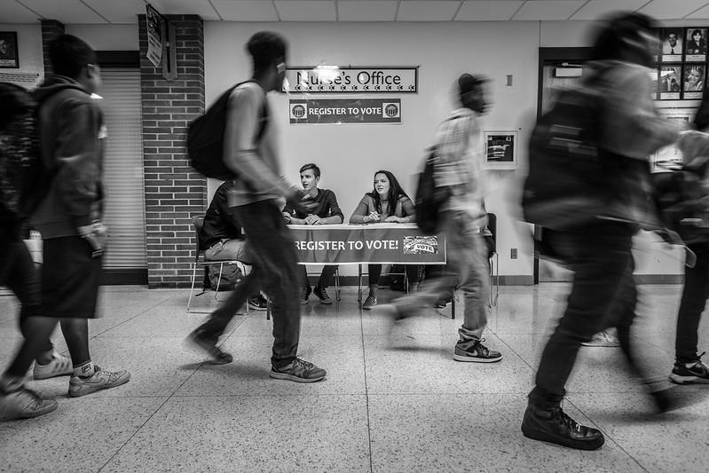 A greyscale photo of a university hallways where students walk in front of a table bearing a 'register to vote' banner