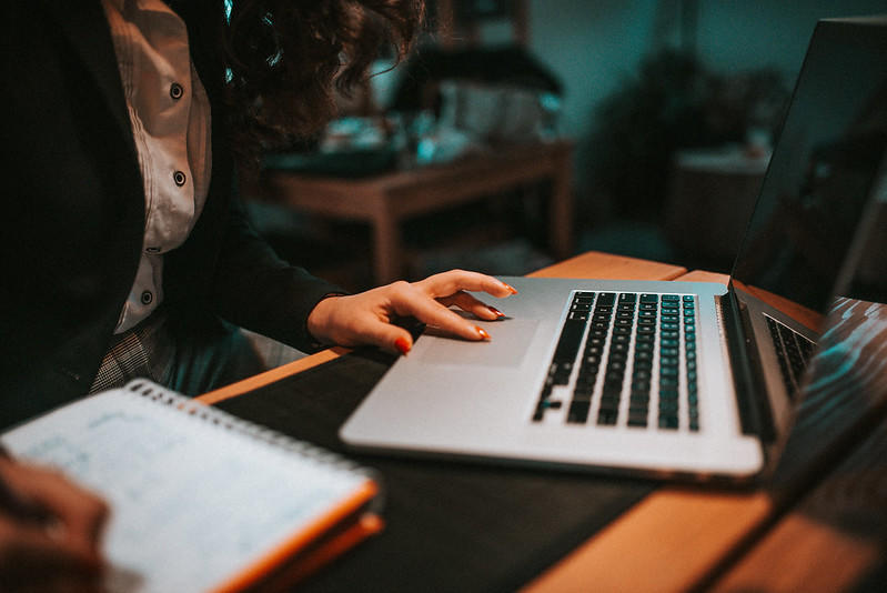 A woman sits at a wooden table in front of an open laptop, working the touchpad while writing in a spiral-bound notebook. 
