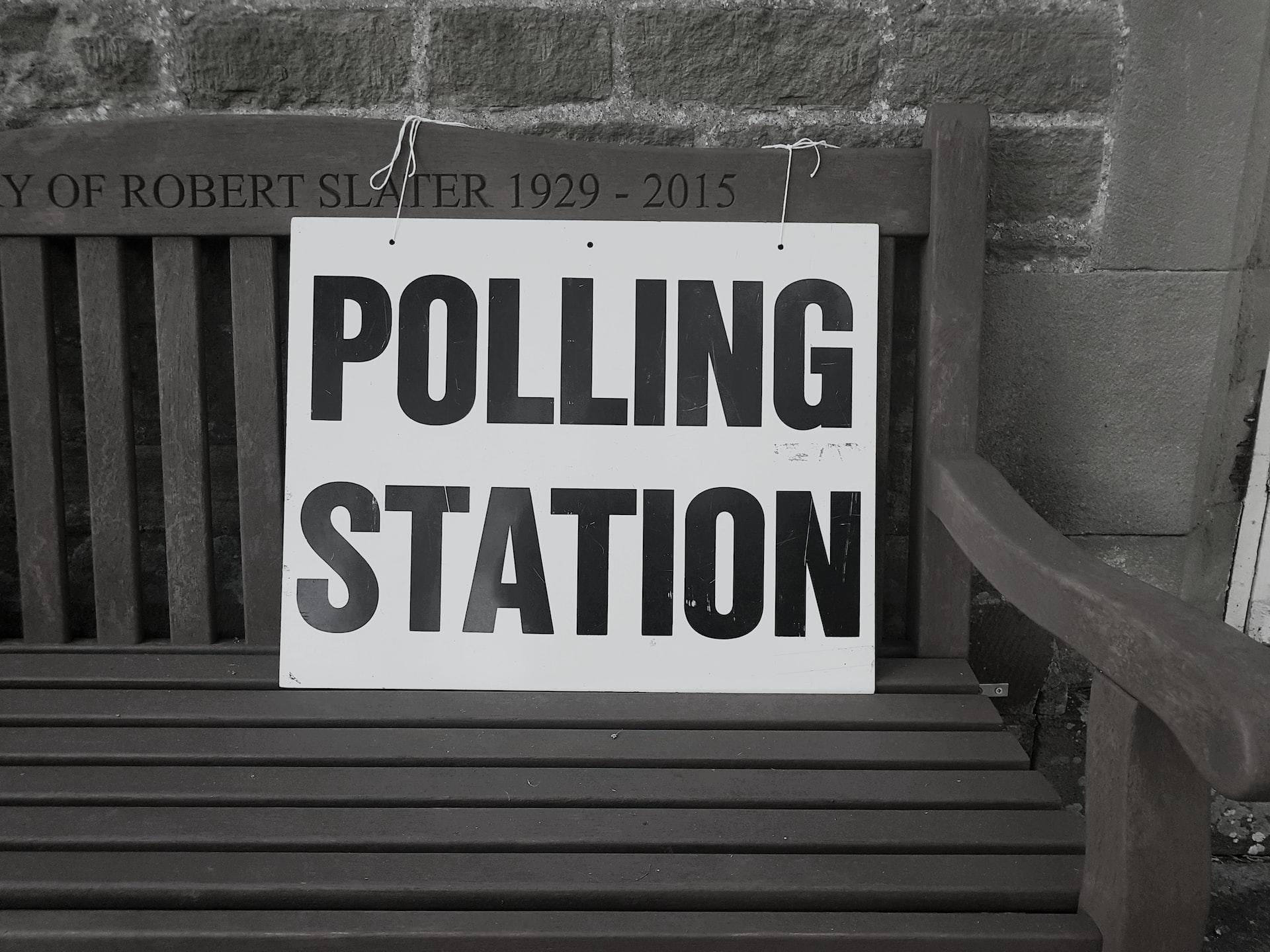 A greyscale picture of a polling station sign propped up on a wooden, slatted bench in front of a brick wall. 