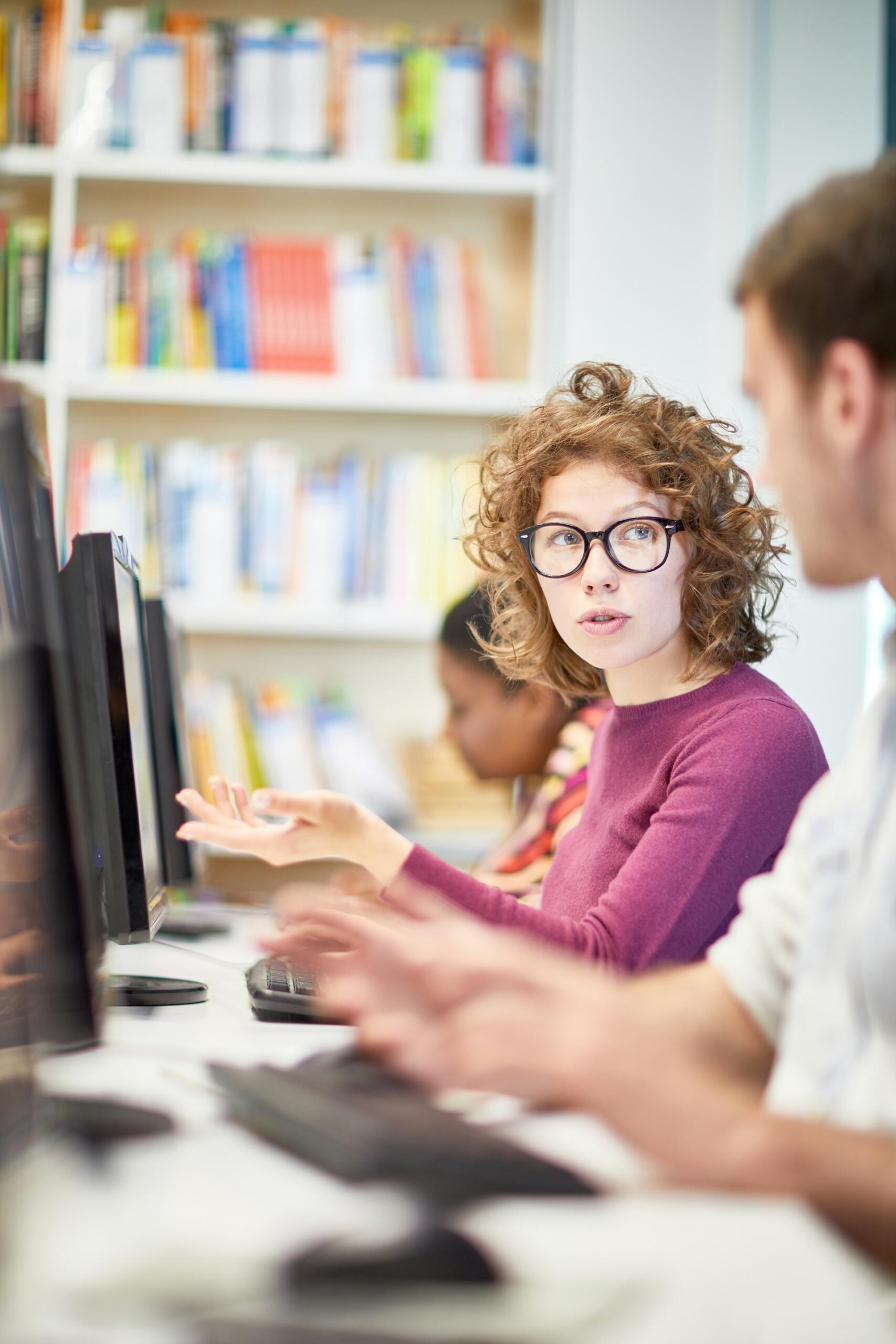 A person in a mauve top wearing glasses asks the person seated next to her a question.