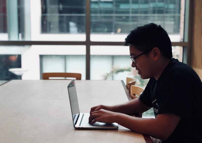 A young man wearing glasses and a black tee-shirt sits alone at a long white table, smiling as he types on his laptop. 