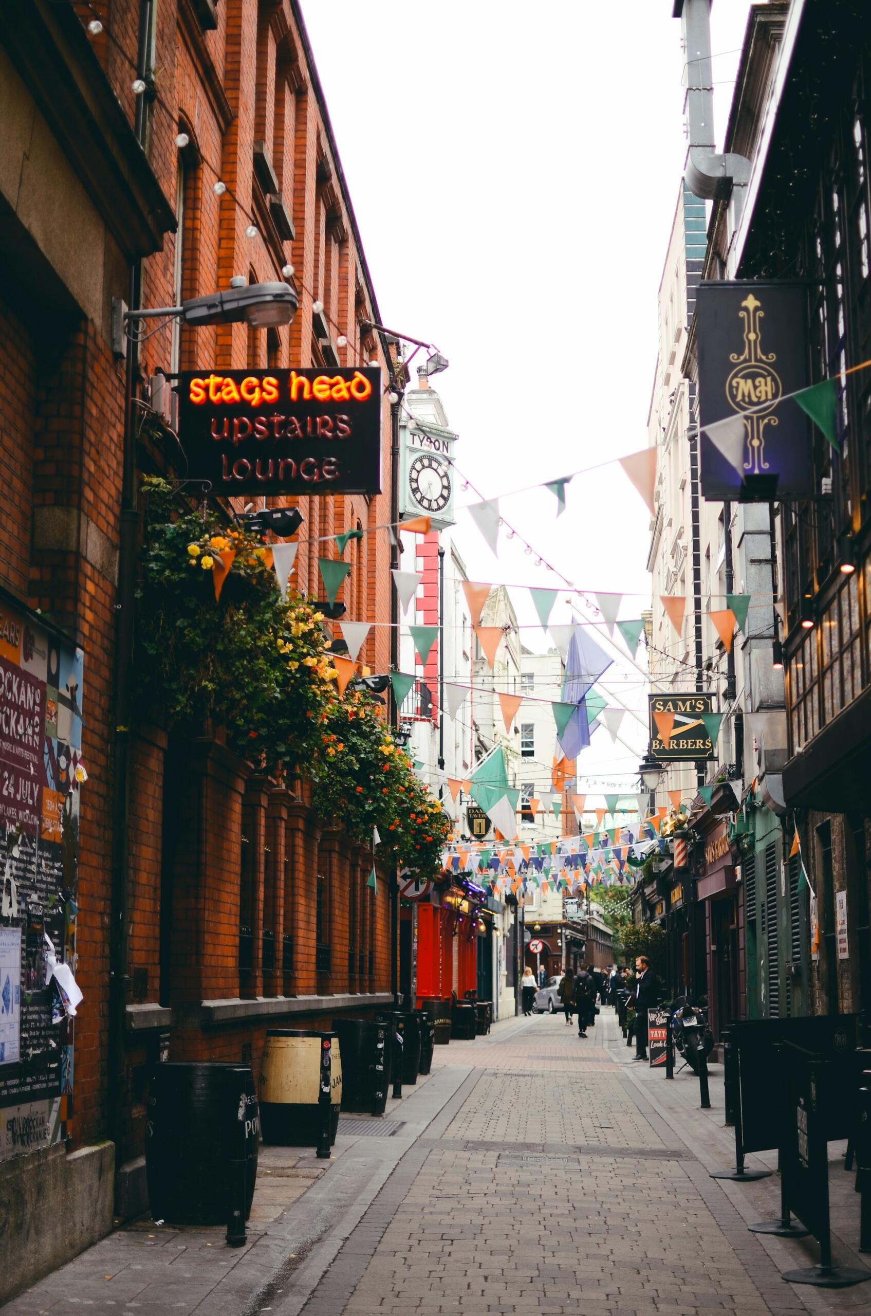 A narrow street with tall buildings, and pennants strung across them.