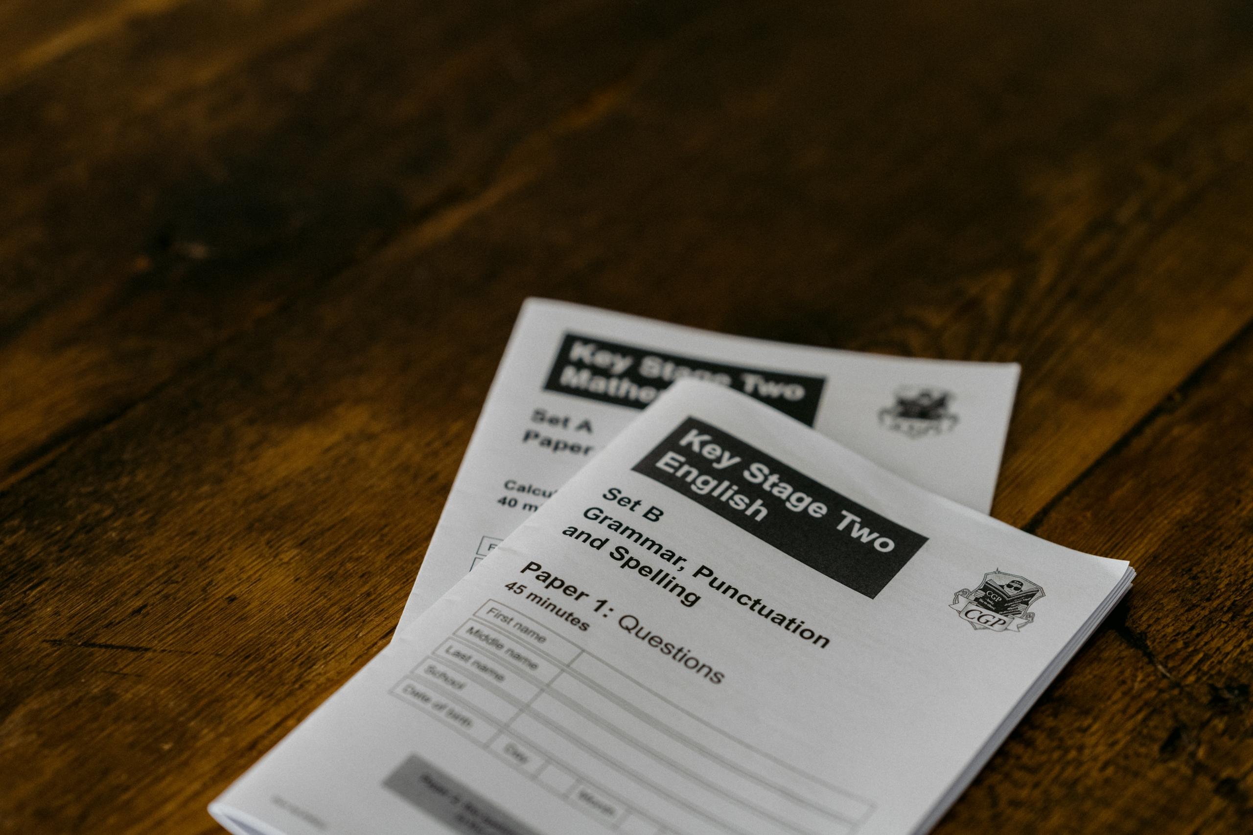 Two Key Stage Two English test booklets laying one atop the other, slightly fanned out, on a dark wooden plank surface. 