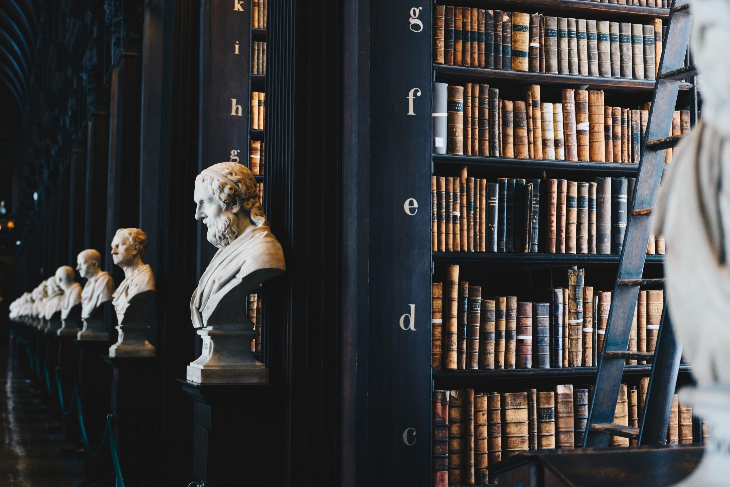 A formal library with seemingly antique hardback books stacked in dark wood shelves, with each stack prefaced by a bust of a renowned person. 