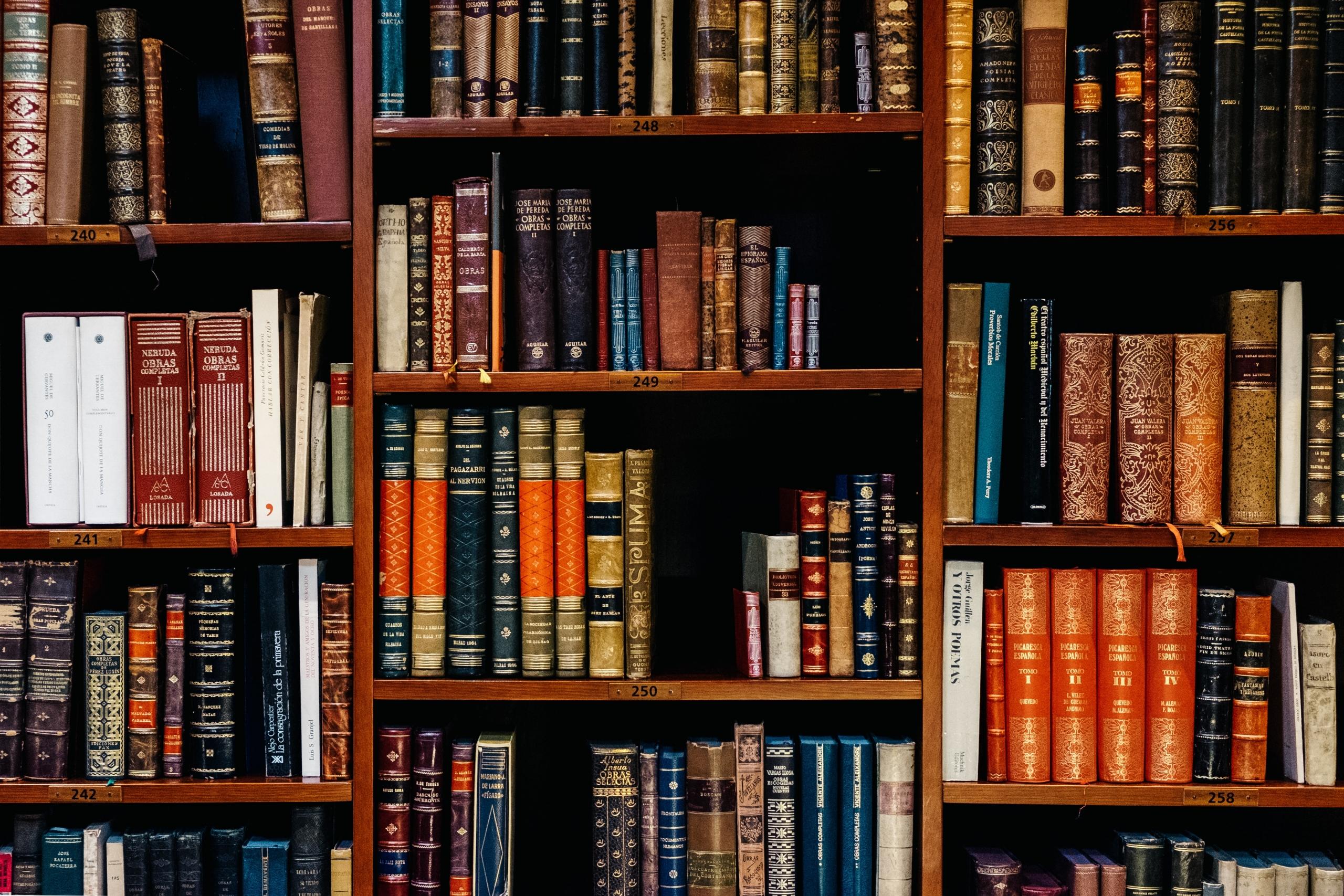 A dark wood book shelf laden with hardback books of various sizes and colours.