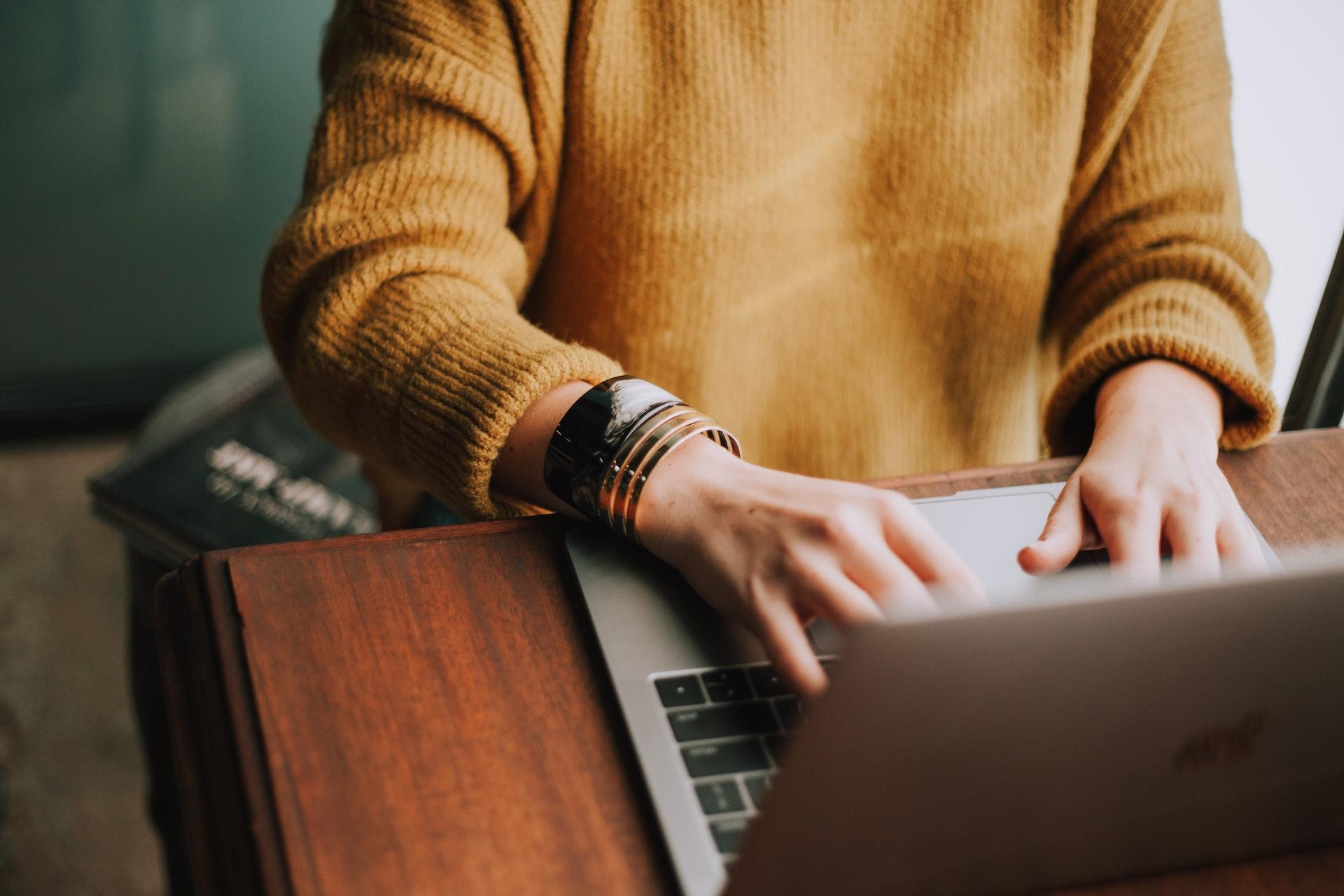 A person with bangles on their wrist in an ochre jumper types on an open laptop computer which sits on a wooden surface. 