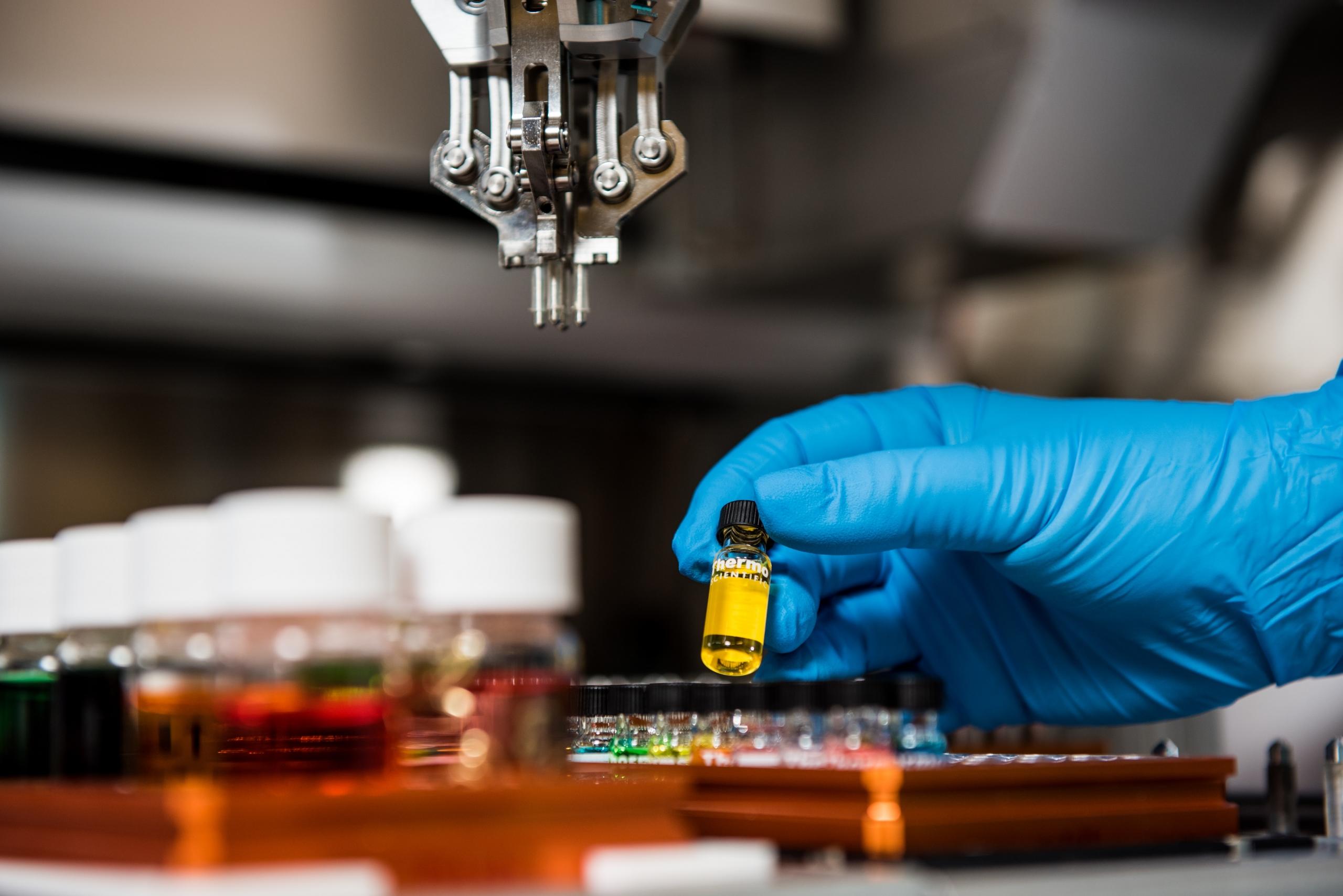 A hand covered with a blue nitrile glove hold a sample vial with a yellow label and black lid above a rack of similar containers.