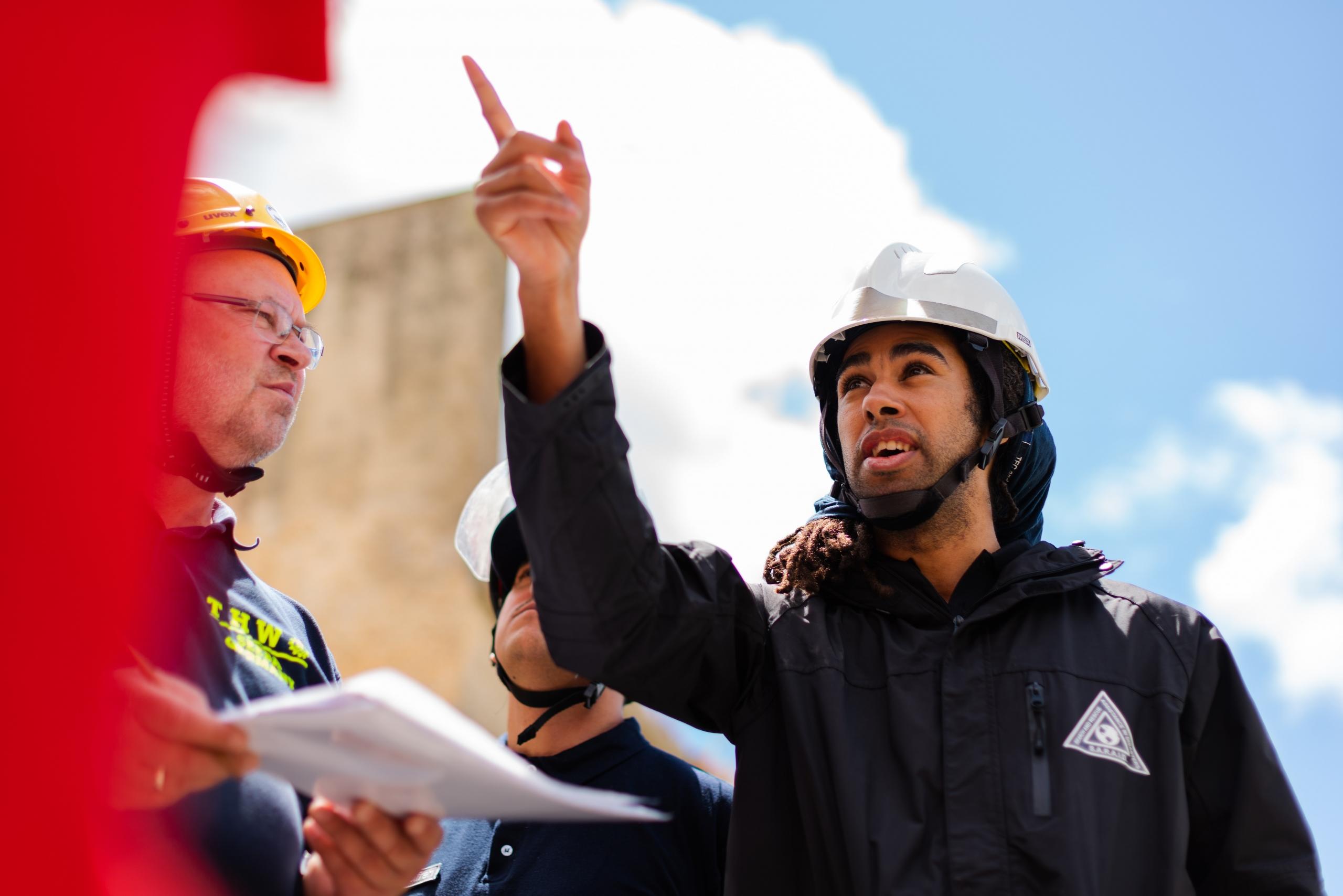 A person wearing a white hard hat with its chin strap fastened and a dark jacket featuring a white triangle over the breast pocket points at something on a red structure as another man wearing glasses and a yellow hardhat looks at him and holds sheets of paper. 