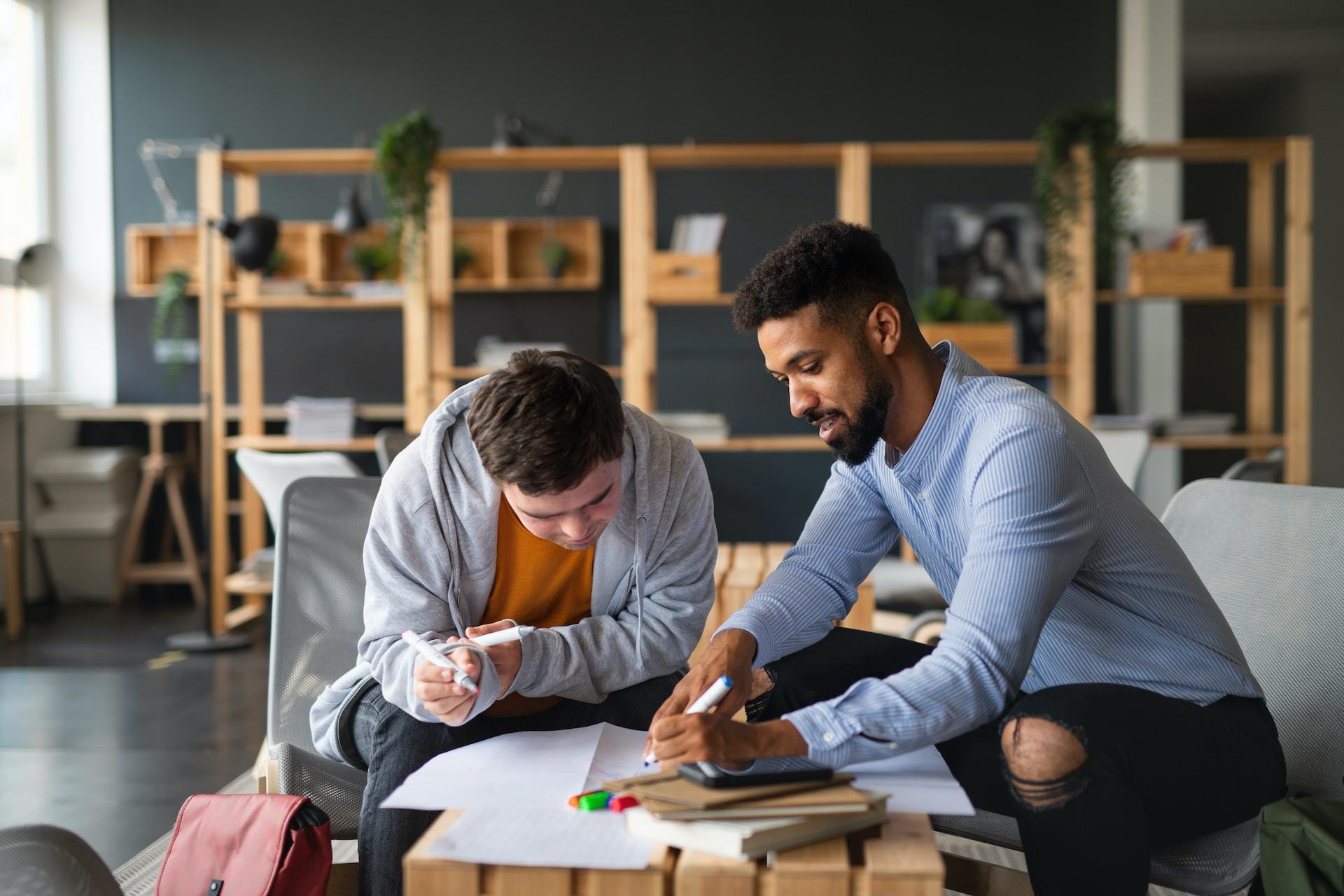 A tutor in a blue shirt holds a pen in their left hand as they write while a student looks on.