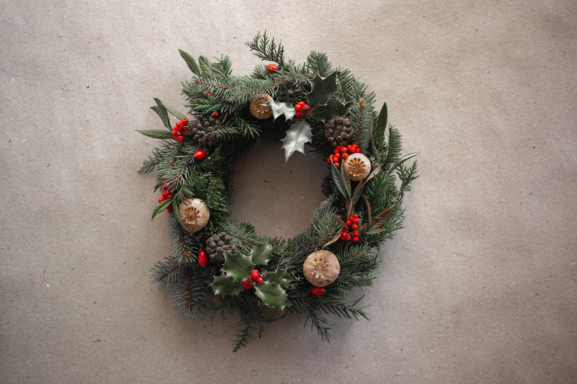 A Christmas wreath made of pine branches, featuring red yew berries and other ornaments, handing on an off-white wall. 