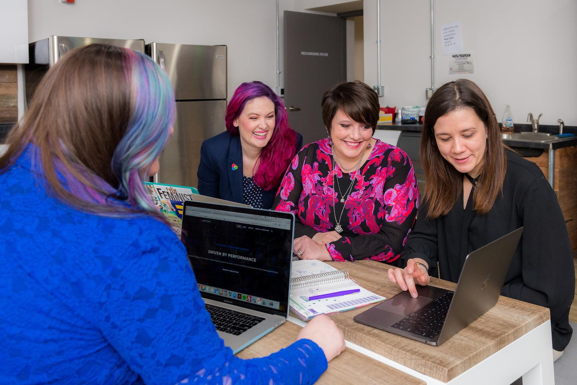 Four women sit at a table, three on one side and one on the other. The table holds two open laptop computers and some papers. The three women on one side look at one laptop while smiling. The fourth woman has an open laptop in front of her.
